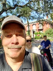 A person is taking a selfie with two other individuals on bicycles in the background, in what appears to be a sunny outdoor setting with trees and a red building.