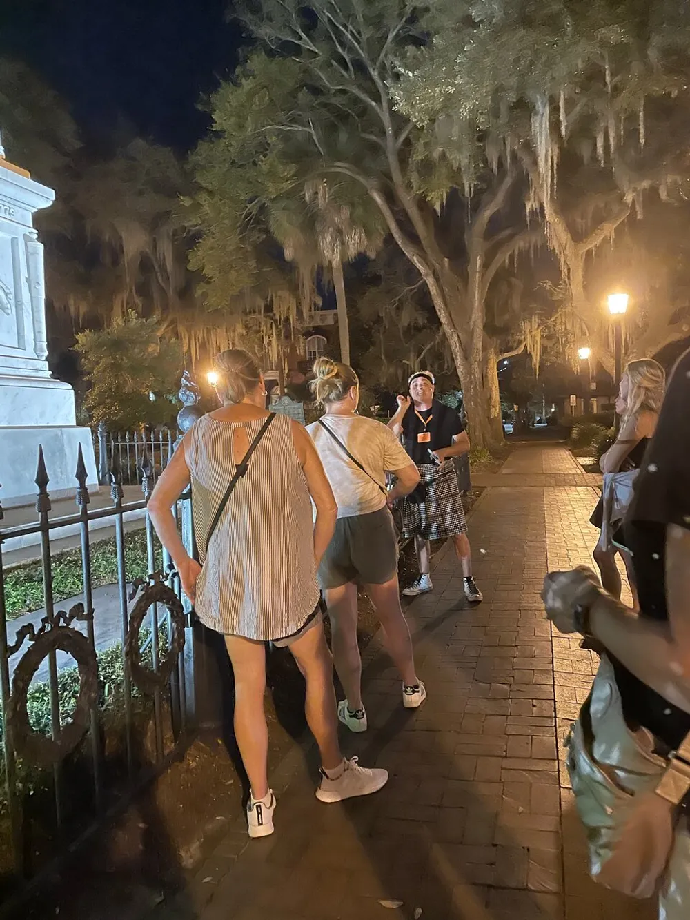 A group of people is gathered at night possibly on a tour listening to a guide who is speaking and gesturing surrounded by trees with Spanish moss and the glow of streetlights