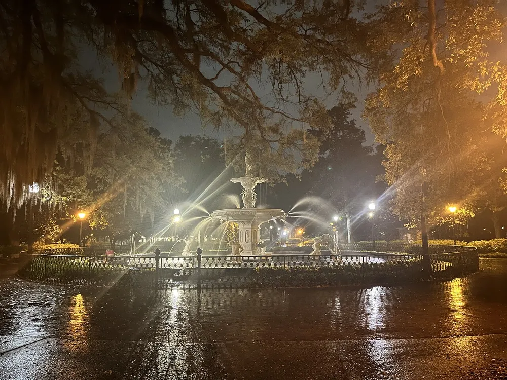 An ornate fountain is illuminated at night by surrounding streetlights creating reflective surfaces on wet ground amidst a setting of hanging Spanish moss