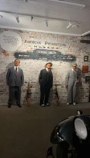 The image appears to be an exhibit at the American Prohibition Museum showing a criminal lineup featuring models or mannequins representing notable figures from the Prohibition era, accompanied by placards with names like Bugs Moran, Al Capone, and Machine Gun Jack McGurn, presented in front of a vintage car's headlight.