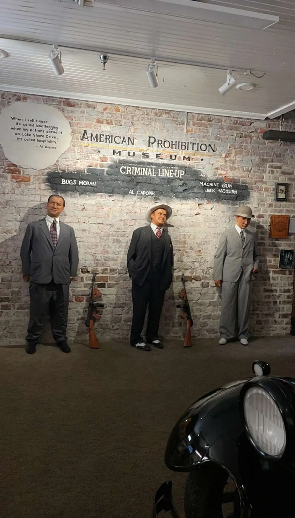 The image appears to be an exhibit at the American Prohibition Museum showing a criminal lineup featuring models or mannequins representing notable figures from the Prohibition era accompanied by placards with names like Bugs Moran Al Capone and Machine Gun Jack McGurn presented in front of a vintage cars headlight