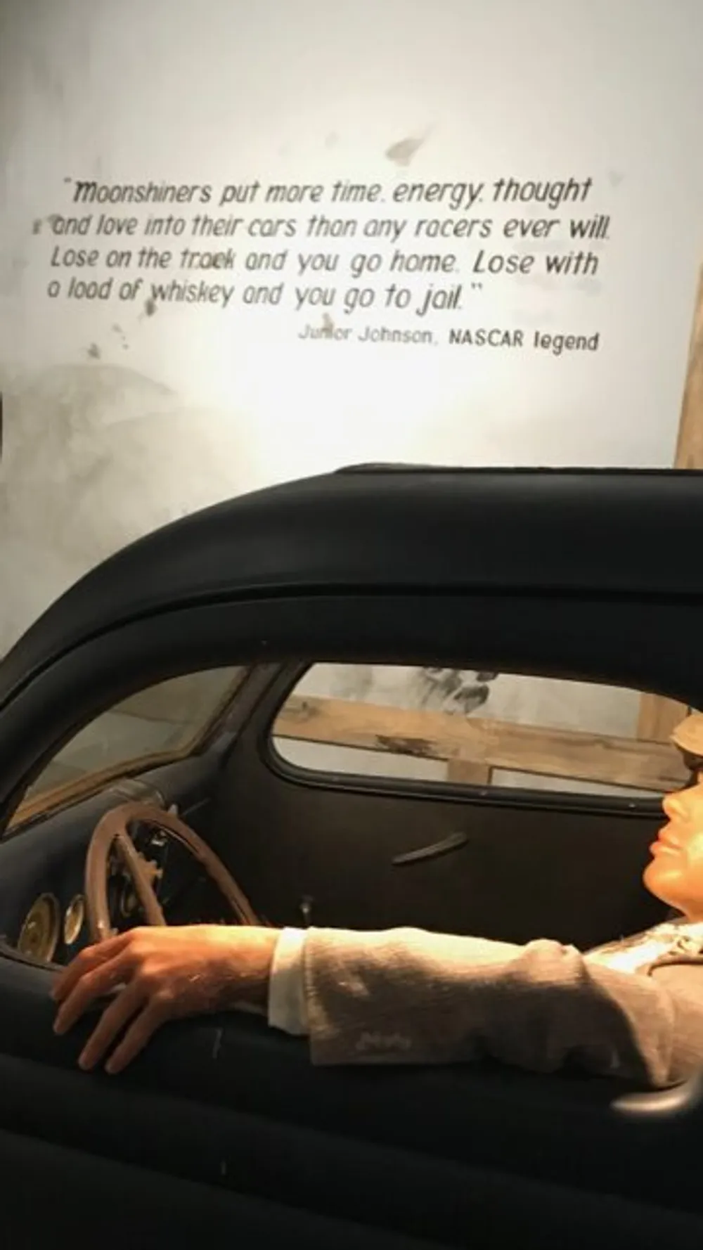 The image displays a mannequin portraying a driver sitting in an old car with a quote from Junior Johnson a NASCAR legend about moonshiners dedication to their cars written on the wall behind