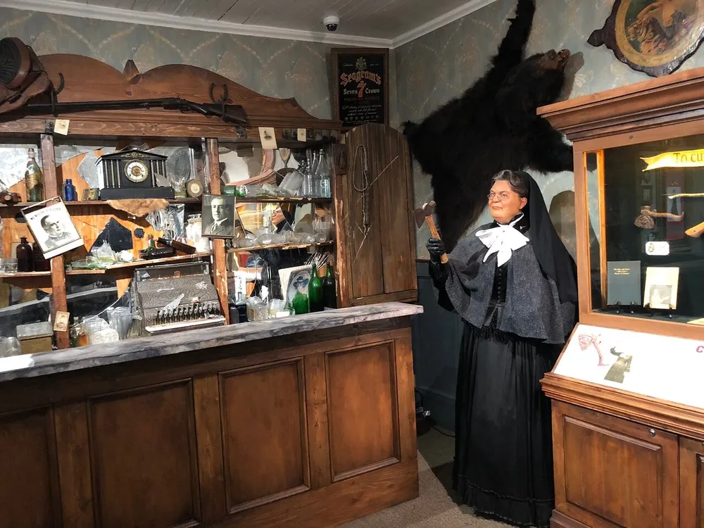 The image shows a vintage-style room with an antique counter a plethora of old-fashioned artifacts on the shelves and a wax figure dressed in period clothing holding a gavel with a taxidermied bear mounted on the wall behind it
