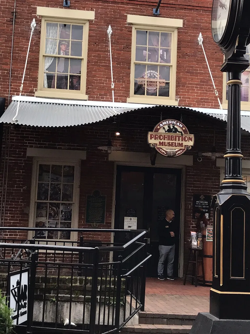 The image shows the exterior of the American Prohibition Museum with a person standing at the entrance under an awning featuring the museums sign set against a red brick building