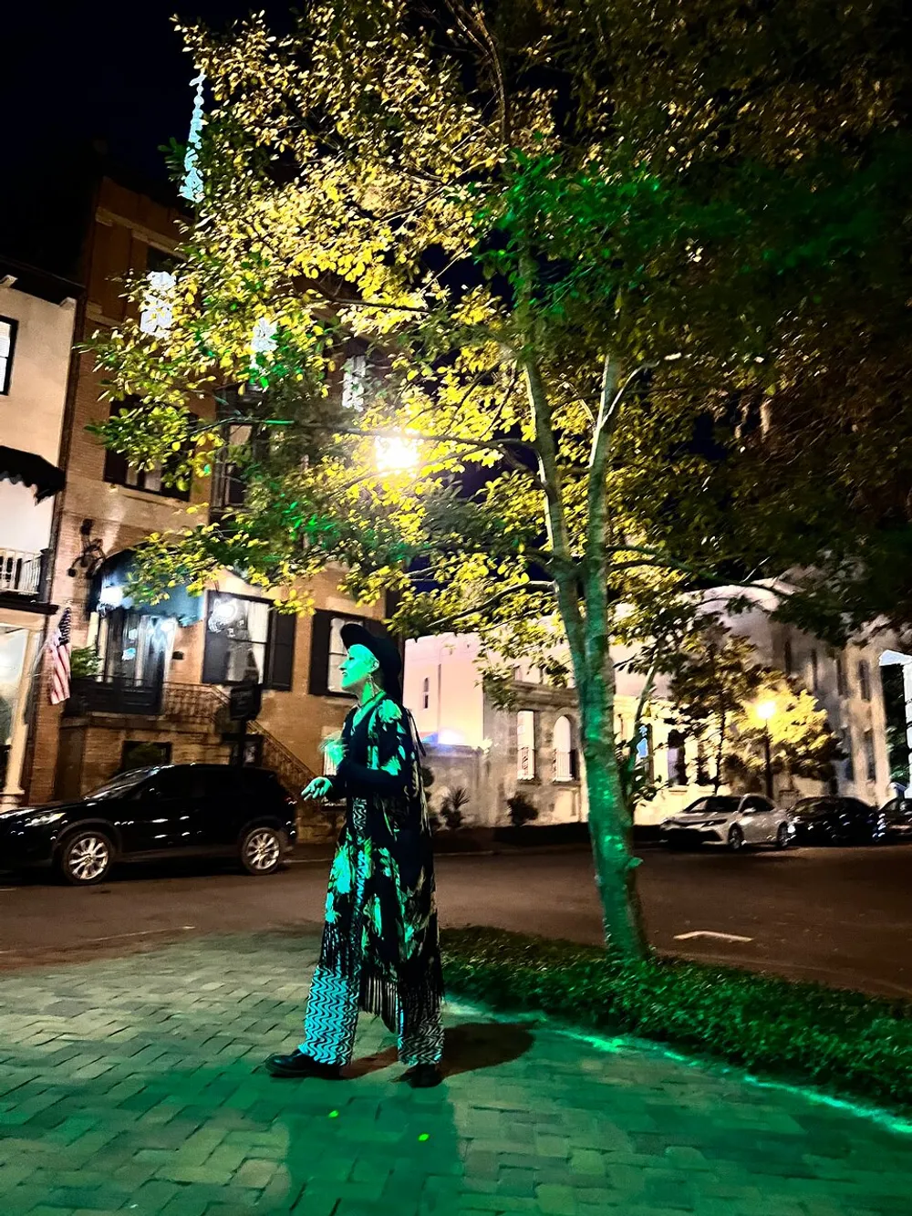 A person stands bathed in green light on a city street at night with a backdrop of illuminated trees and buildings