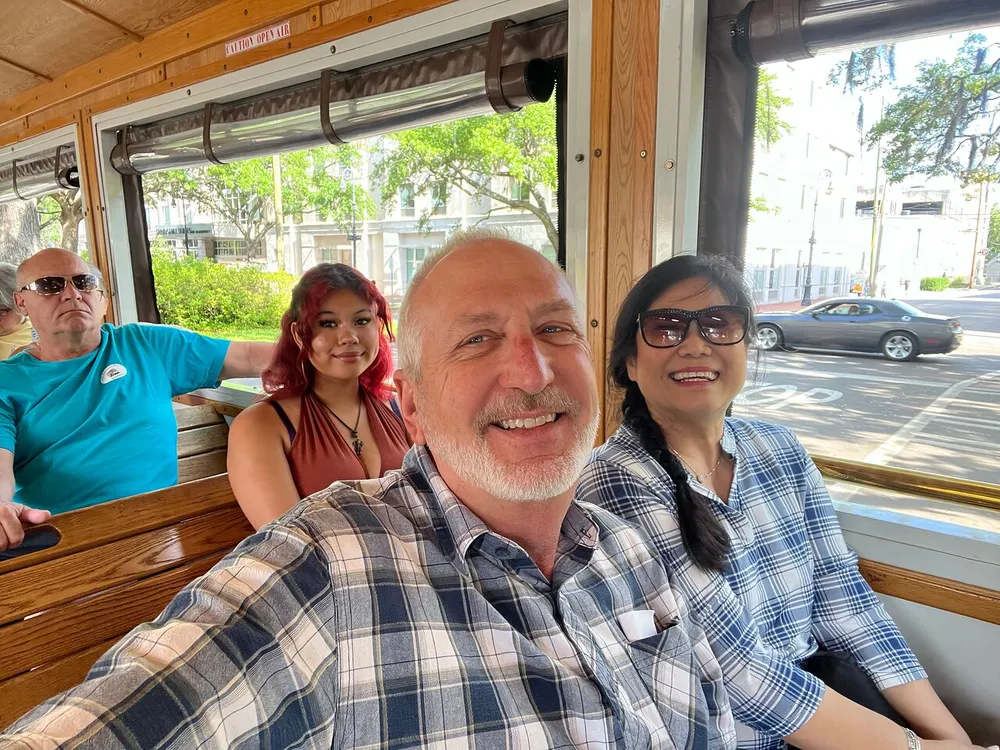 A group of people is enjoying a ride on a trolley with a man taking a selfie including himself and two women sitting beside him and another man in the background