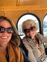 Two women wearing sunglasses are smiling for a selfie inside a vehicle with wooden interior details.
