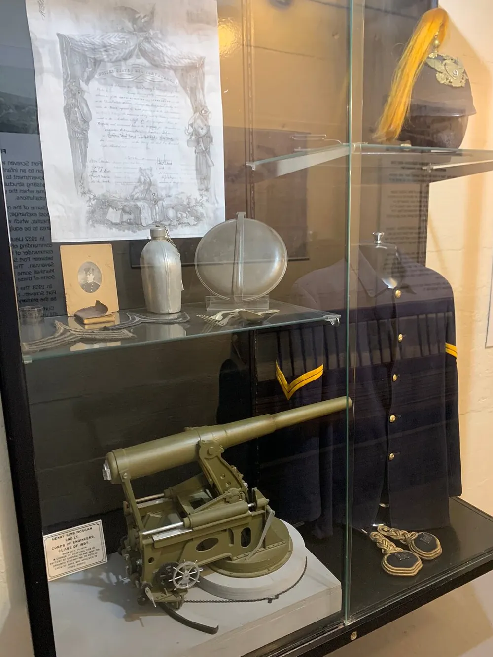 The image shows a display case containing military memorabilia including a model of a heavy machine gun a canteen uniform with rank insignia a helmet with a plume and other items likely part of a historical exhibit