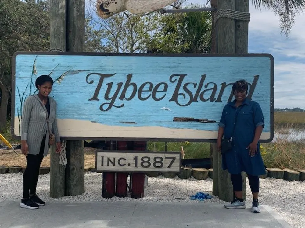 Two individuals are posing in front of a weathered Tybee Island sign which indicates the location was incorporated in 1887