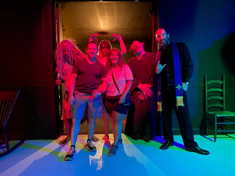 A group of people is posing on a stage with eerie lighting and two individuals looking like they are part of a spooky or Halloween-themed performance