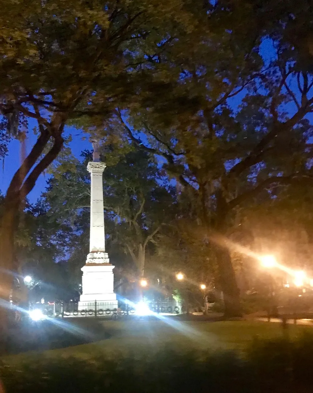 The image depicts a nighttime view of a tall monument illuminated by artificial lights and surrounded by trees with visible lens flare from the lights