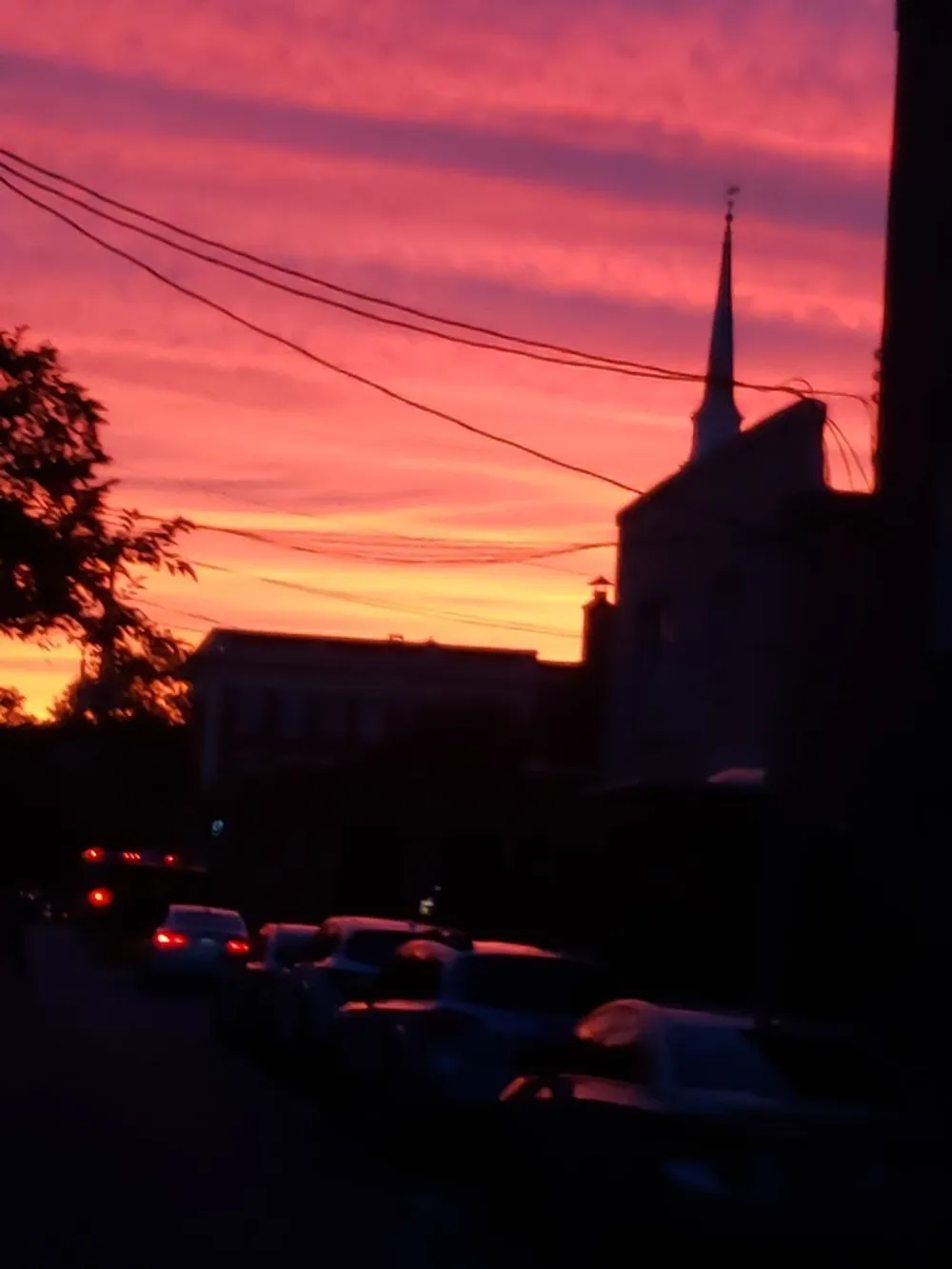 The image captures a vibrant sunset sky in shades of pink and orange above a silhouette of a streetscape featuring buildings a church spire and parked cars