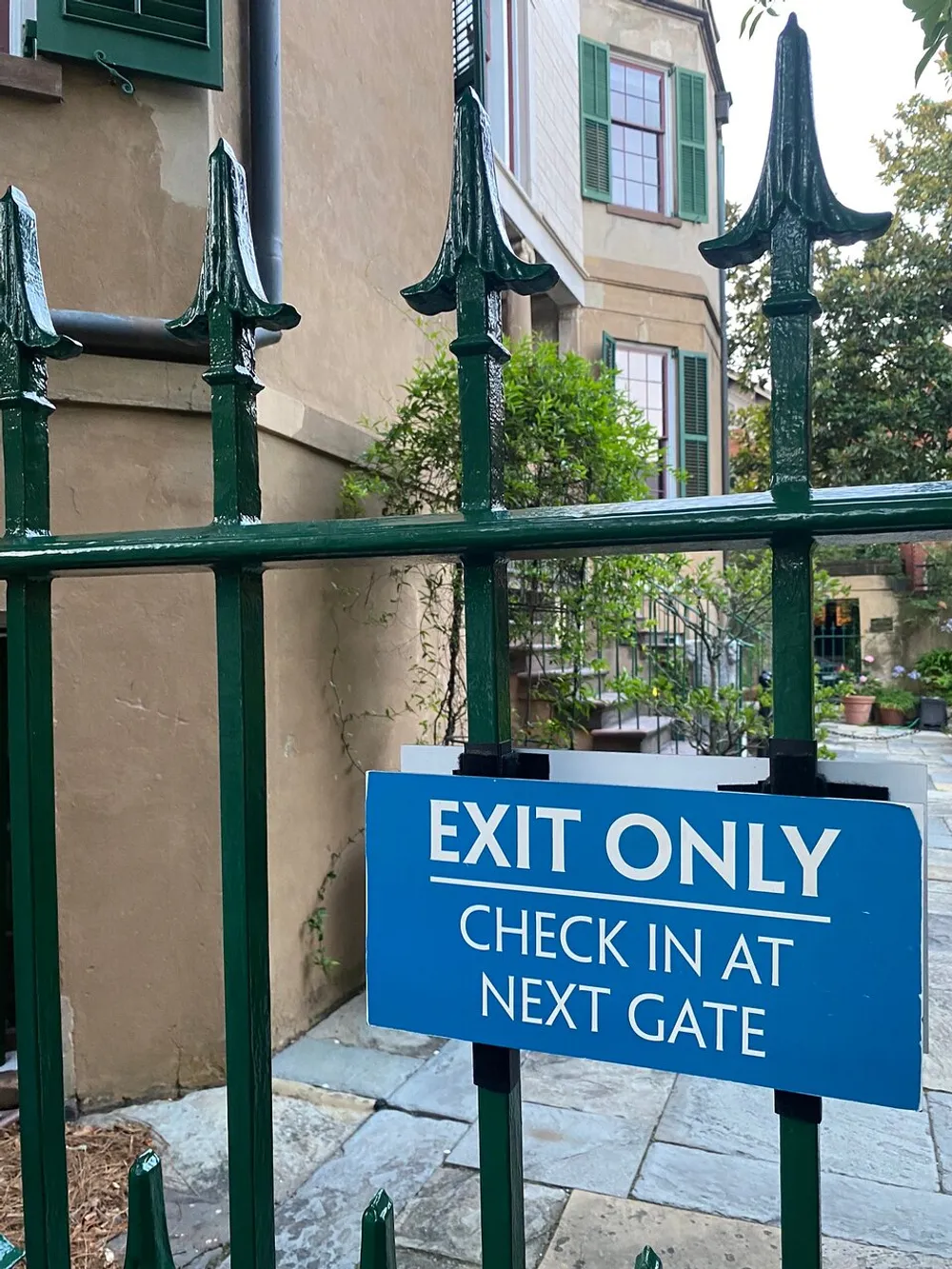 The image shows a blue sign attached to a green metal fence with pointed tips that reads EXIT ONLY CHECK IN AT NEXT GATE indicating directional flow for entrance or exit against a background featuring a portion of a building with green shutters