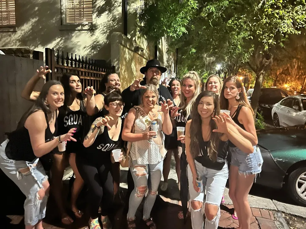 A group of people mostly women wearing black SQUA tank tops are posing for a photo at night with playful expressions along with a man in a black hat at the center in what appears to be a lively outdoor setting
