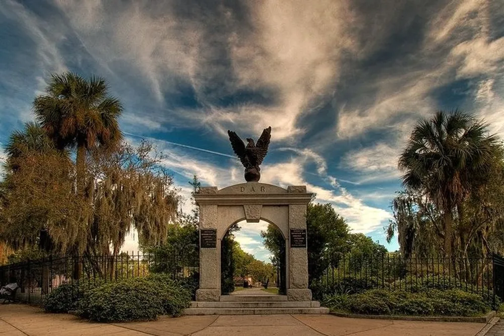 An impressive eagle statue perches atop an archway inscribed with DAR surrounded by lush greenery under a dynamic sky