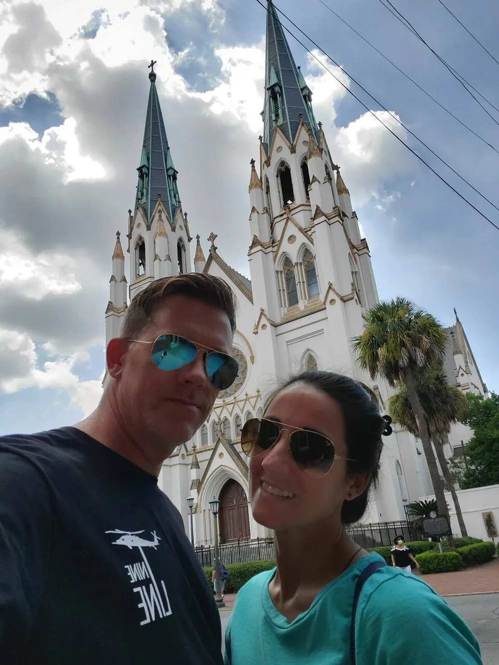 A man and a woman are taking a selfie with an ornate church featuring twin spires and Gothic architecture in the background