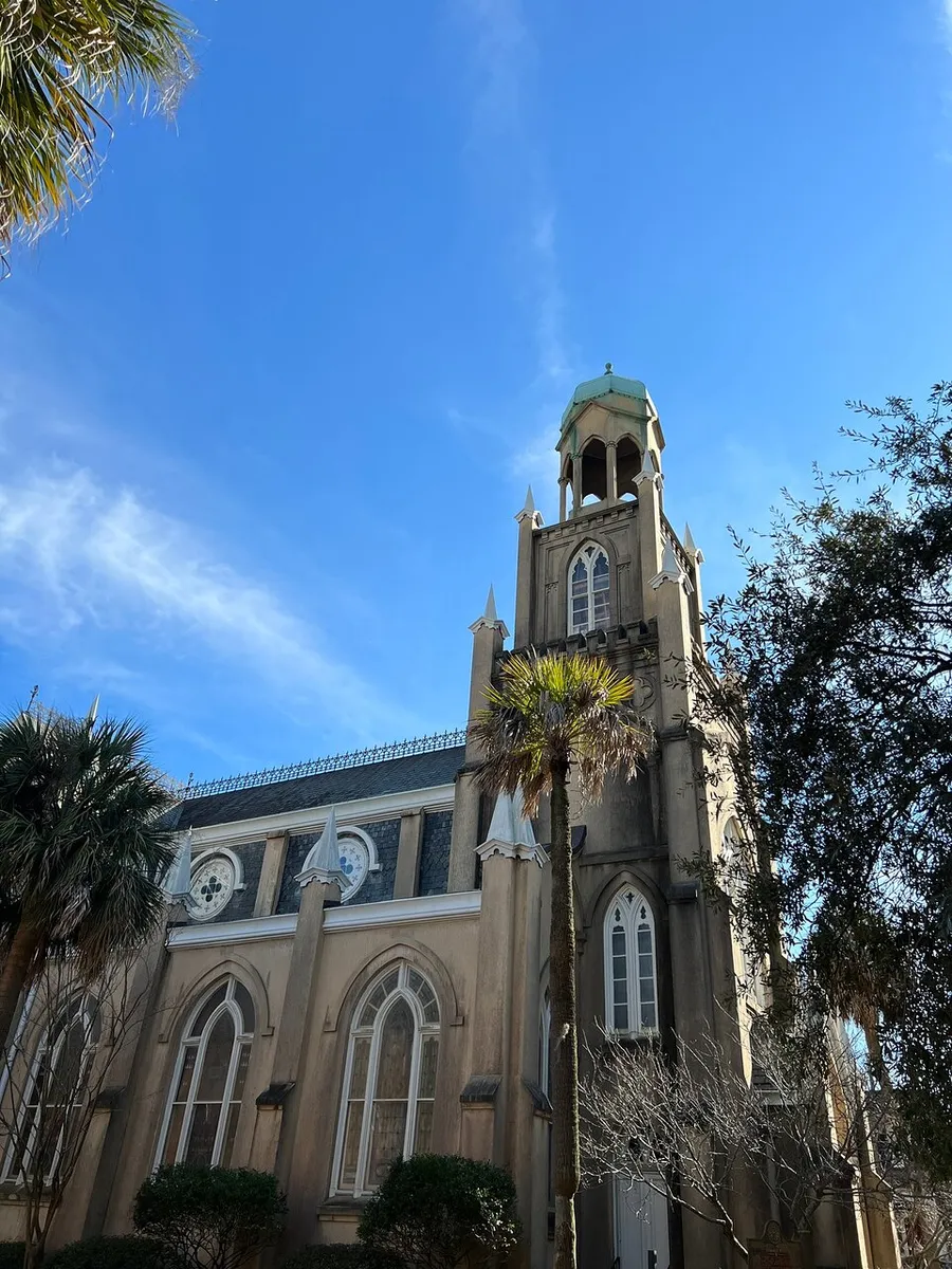 The image shows a tall, historic-looking church with gothic architectural elements, framed by palm trees against a clear blue sky.
