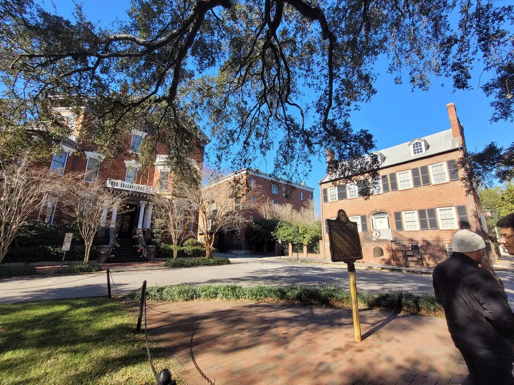 The image depicts a sunny day at a location with historic-looking brick buildings where a few individuals are walking and the scene is framed by mature trees