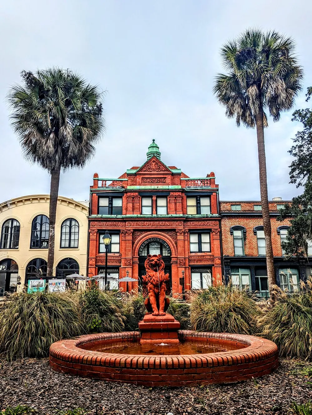 The image shows a picturesque view of a historic red brick building with the inscription Savannah Cotton Exchange framed by palm trees and a fountain with a lion statue in the foreground