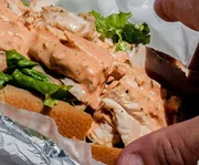 The image shows a close-up view of a chicken sandwich with lettuce and a creamy sauce on a hoagie roll, partially wrapped in foil and being held by a person.
