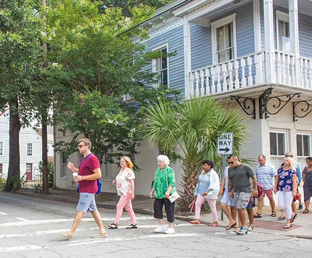A group of people is casually walking down a tree-lined street with historical buildings and a One Way street sign in view