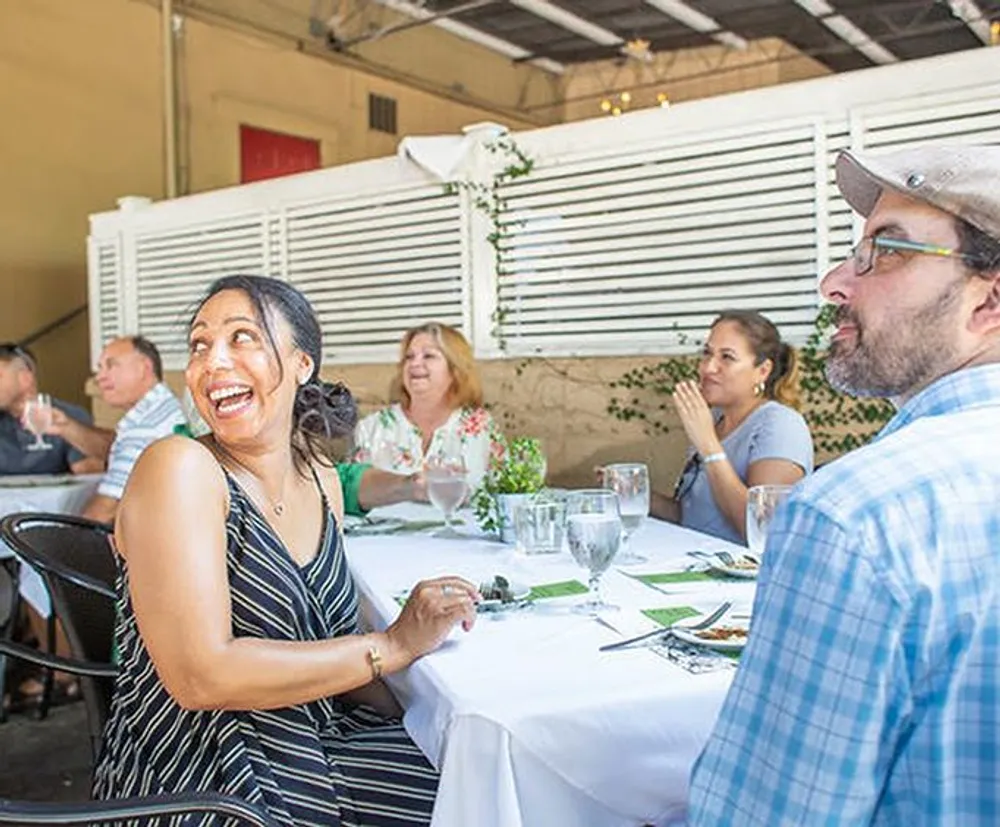A joyful woman is laughing while sitting at a table in an outdoor restaurant setting accompanied by other diners enjoying their meals