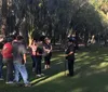 A group of people is standing outdoors in a park or square likely listening to a guide or speaker near a statue and a flowerbed