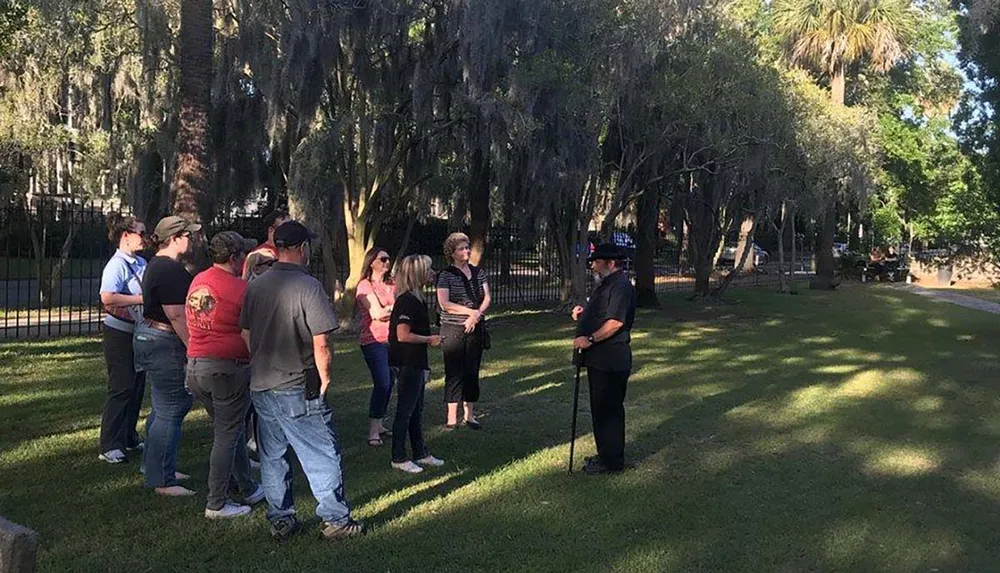 A group of people listens attentively to a person dressed in a historical costume in a park shaded by trees draped with Spanish moss