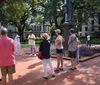A group of people is standing outdoors in a park or square likely listening to a guide or speaker near a statue and a flowerbed