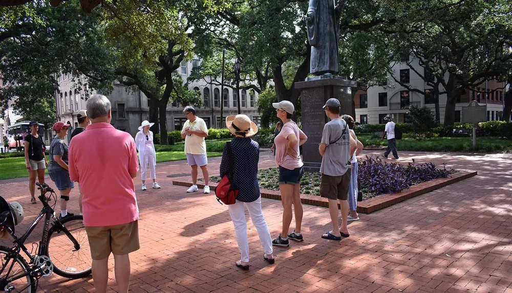 A group of people is standing outdoors in a park or square likely listening to a guide or speaker near a statue and a flowerbed