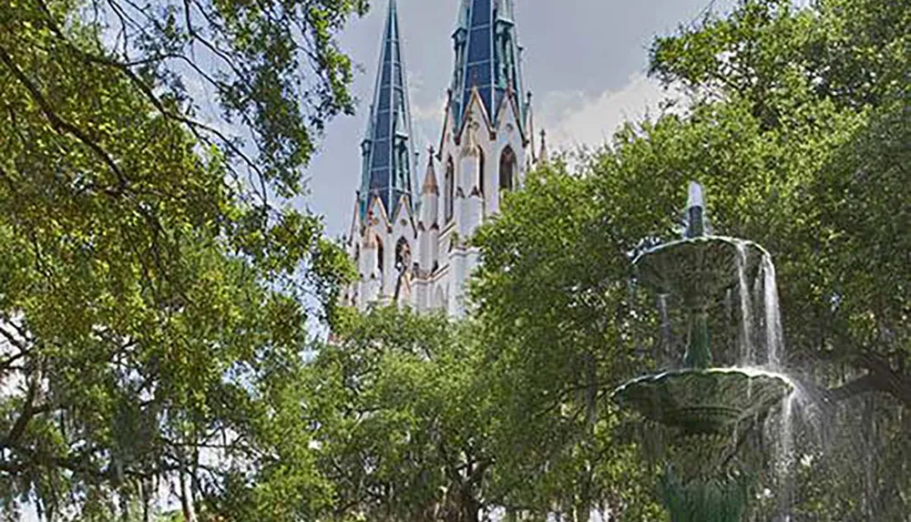 The image features a towering Gothic-style cathedral spire rising behind the lush greens of tree canopies with a classic tiered fountain in the foreground
