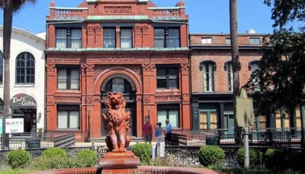 The image depicts a distinguished red brick building with ornate detailing and a large lion sculpture in front situated in a sunny tree-lined urban setting