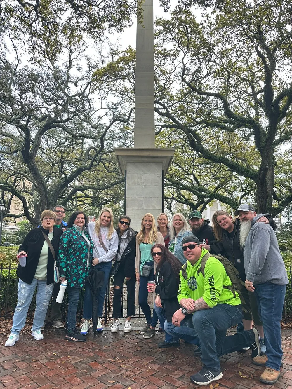 A diverse group of people posing for a photo in a park with a monument and sprawling oak trees in the background