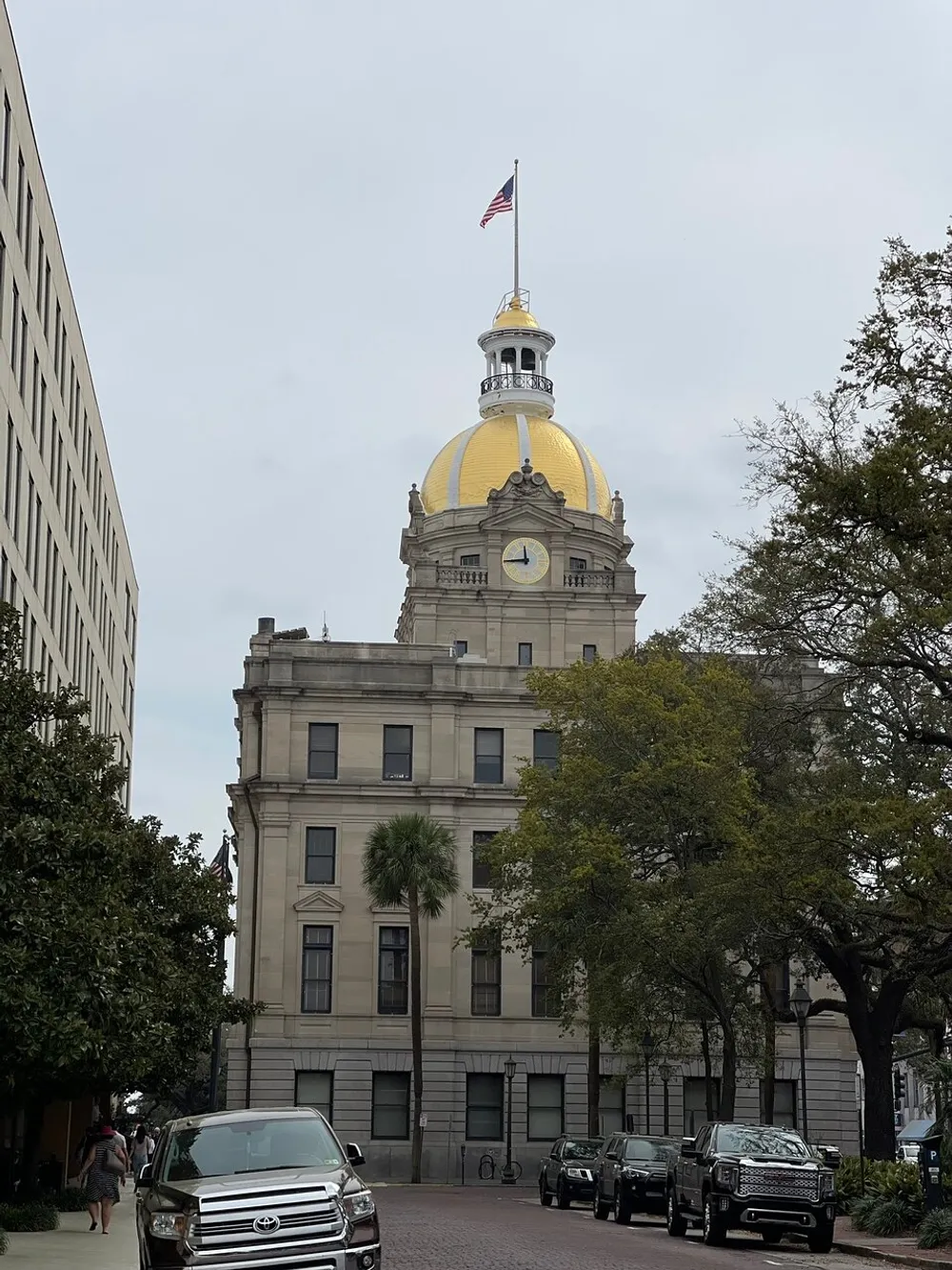 The image shows a street view featuring the dome of a grand historic building with an American flag on top flanked by modern office buildings and vehicles parked along the curb under an overcast sky