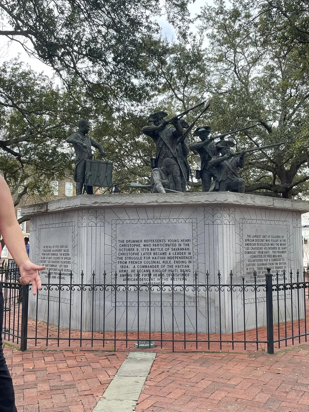 The image shows a monument with figures of historical significance along with a descriptive plaque and a partial view of a persons arm and hand set against a backdrop of trees and brick pavement