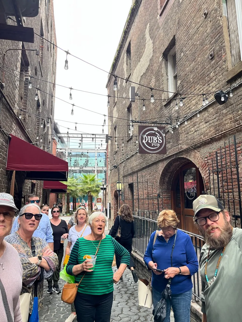 A group of people are standing in a narrow cobblestone alleyway with string lights overhead a pub sign visible and a mix of expressions suggesting they are engaged in a tour or group event