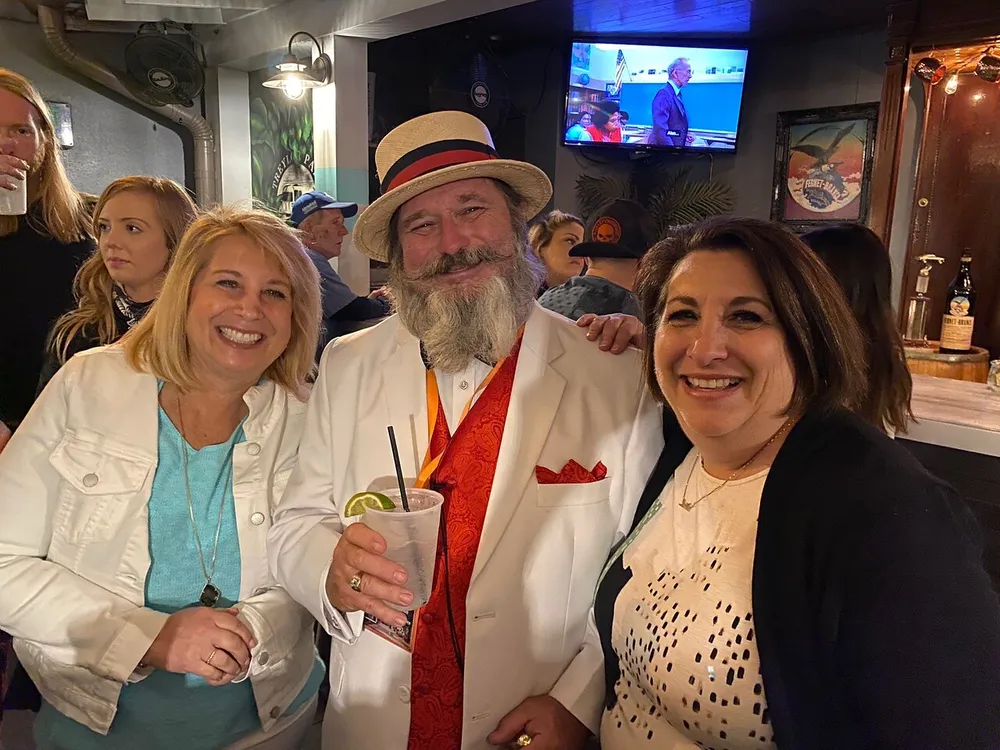 A man in a straw hat and white suit with a red pocket square is smiling with two women by his side in a social setting that appears to be a bar or pub