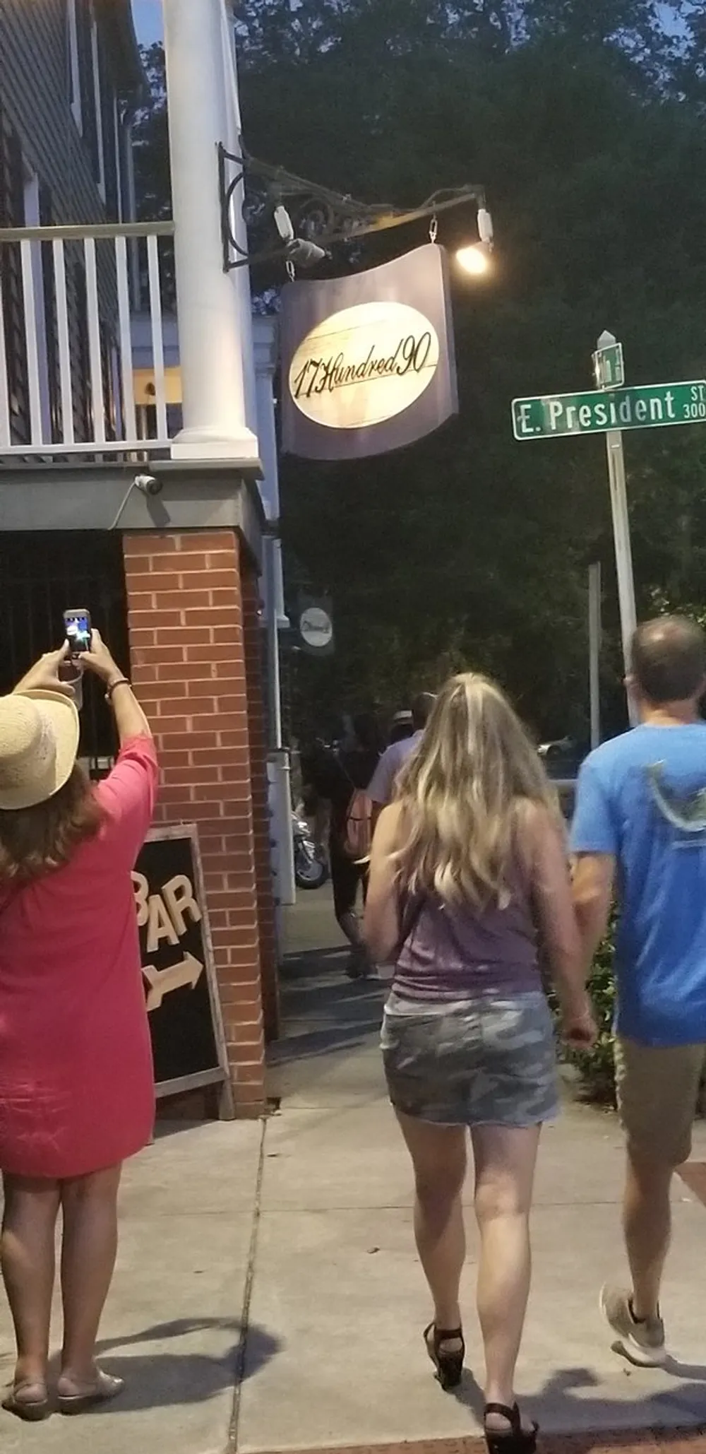Pedestrians are walking on a sidewalk at dusk past a building with a sign that says 17Hundred90 and a woman is taking a photo with her smartphone