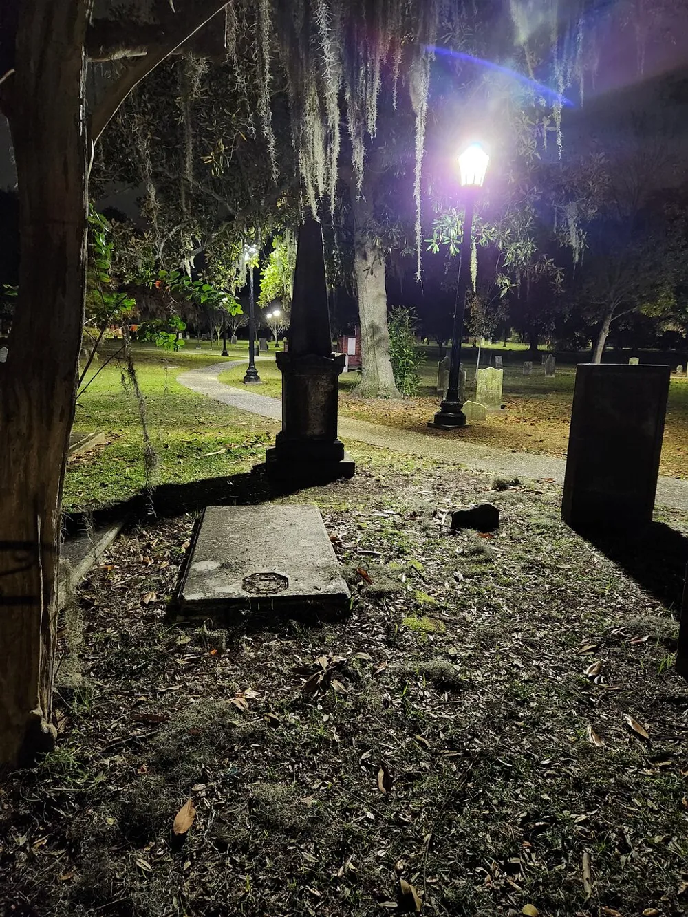 The image shows a serene nocturnal scene of a cemetery with a lit lamp post moss-laden trees and tombstones creating a somber and tranquil atmosphere