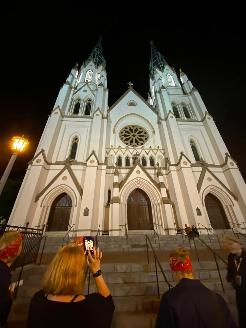 Several people are standing in front of a grand illuminated Gothic-style church at night while one person takes a photo of the intricate facade with their smartphone