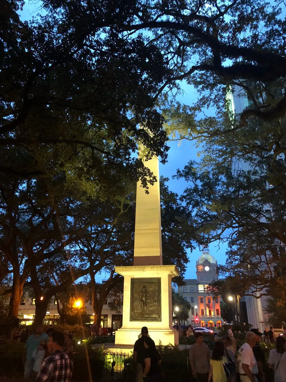 The image presents a bustling evening scene in a park with an obelisk monument surrounded by mature trees and illuminated by ambient street lights with a clock tower in the background