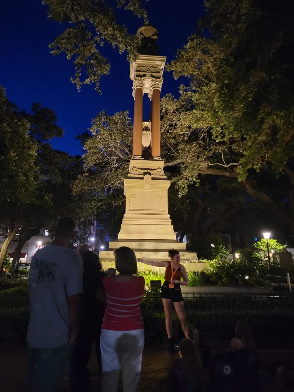 People gather around a tall monument illuminated by ambient light in a park during nighttime