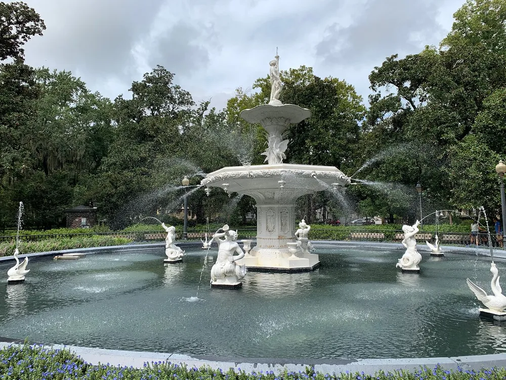 An ornate white tiered fountain with sculptures of swans and children spraying water surrounded by greenery and flowers under a cloudy sky