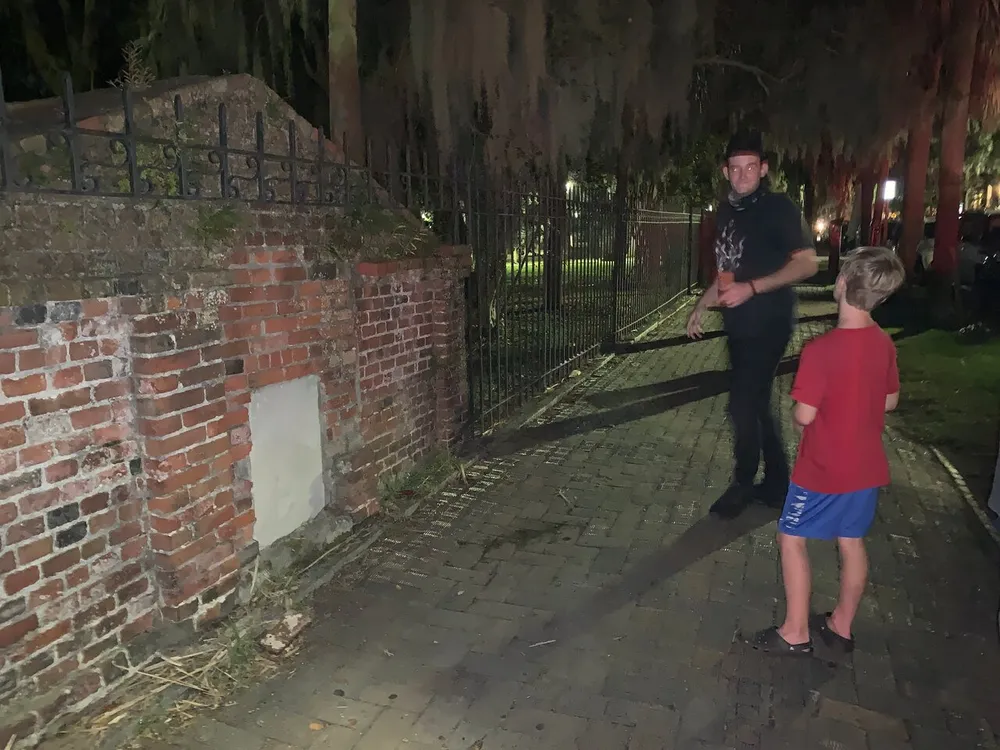 A man and a boy stand near a brick wall and metal fence at night with Spanish moss hanging from trees in the background