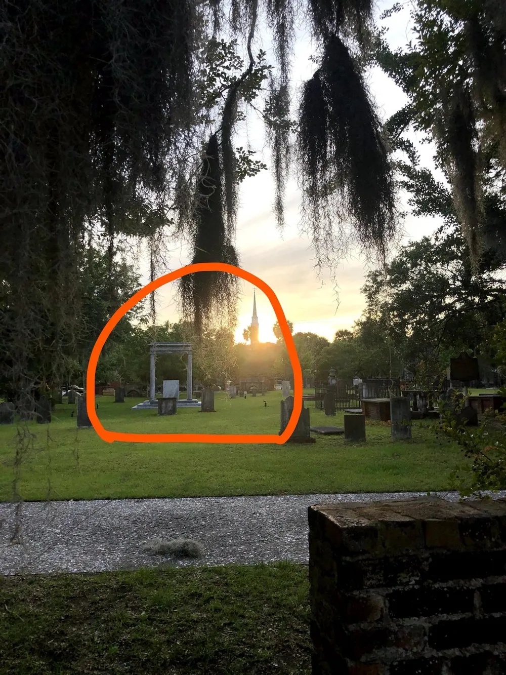 The image captures a serene sunset with golden rays filtering through the trees highlighting a peaceful cemetery surrounded by hanging Spanish moss with an orange outline focusing on a particular area within the scene