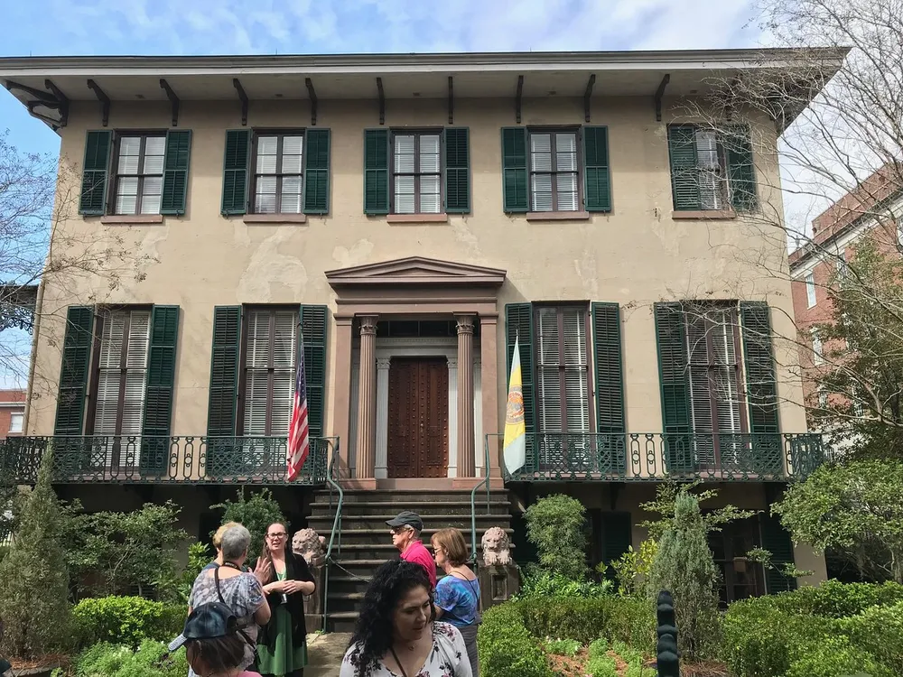 A group of people gather outside a stately two-story building with green shutters and a wrought iron balcony which is adorned with an American flag and another flag