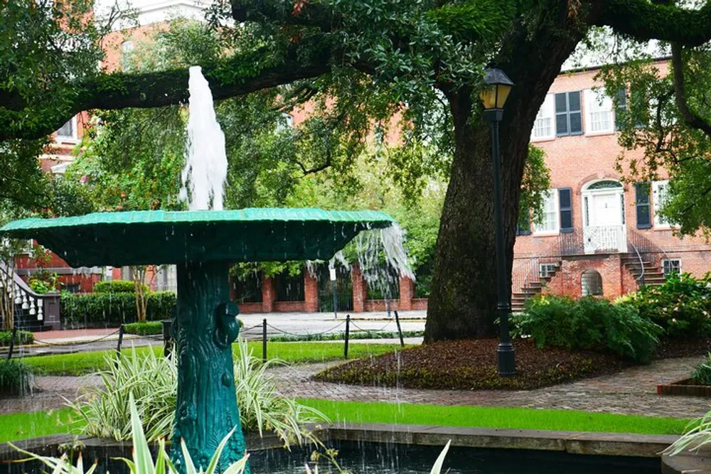 This image shows a green water fountain with water cascading down its tiers surrounded by lush greenery with a lamp post and a red brick building in the background suggesting an urban park setting