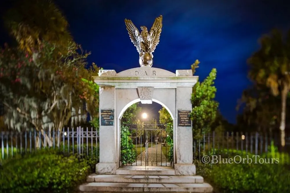The image shows a grand archway entrance at night with a large eagle sculpture on top and the letters DAR engraved leading to what appears to be a path lined with fencing and trees bathed in the glow of a street lamp