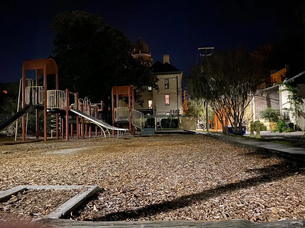 An empty playground is cast in the ambient glow of nighttime lighting framed by the silhouette of urban structures and trees