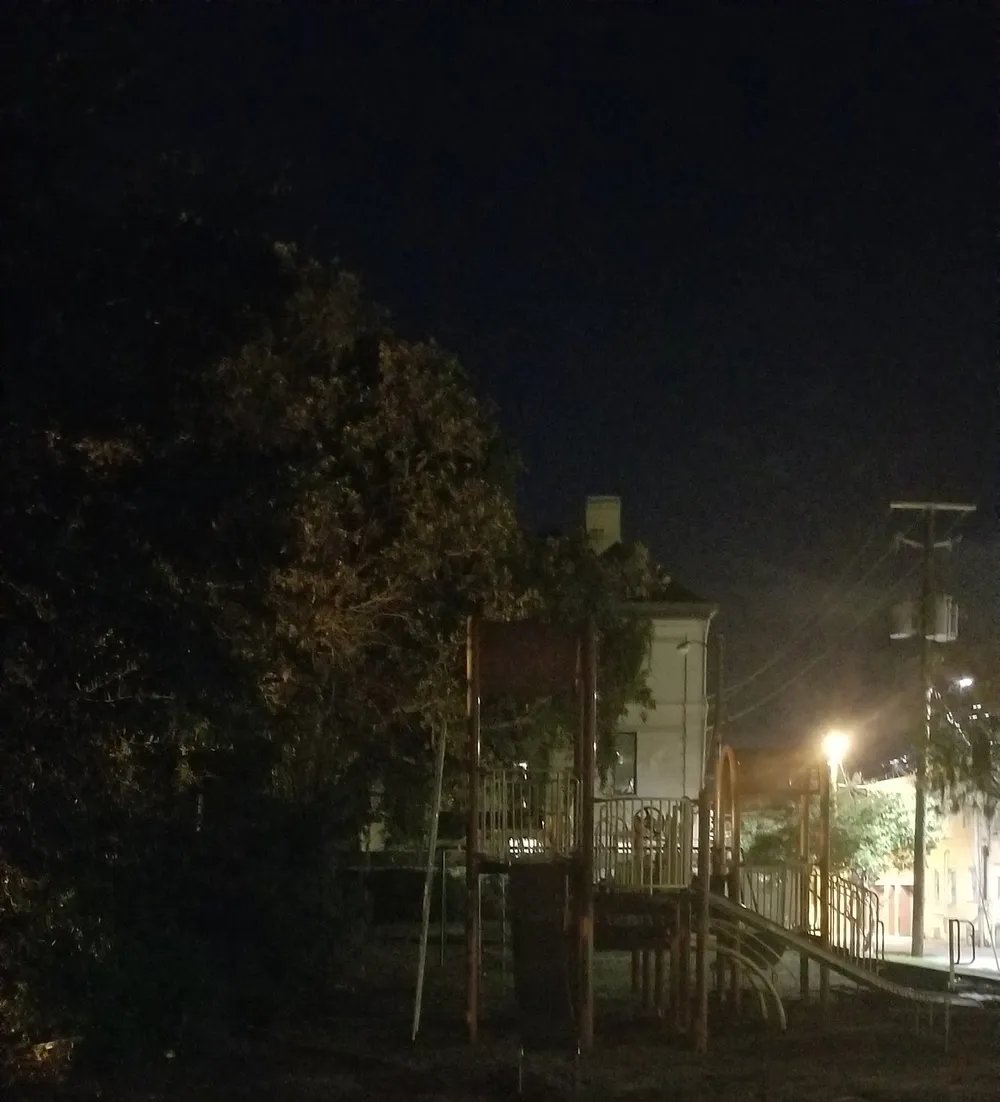 The image shows a dimly lit playground at night with surrounding trees and a building in the background
